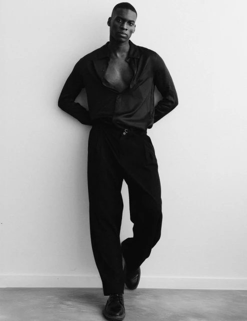 A Black male model poses against a white wall in an unbuttoned black shirt and wrap pants, hands behind back, wearing black shoes.