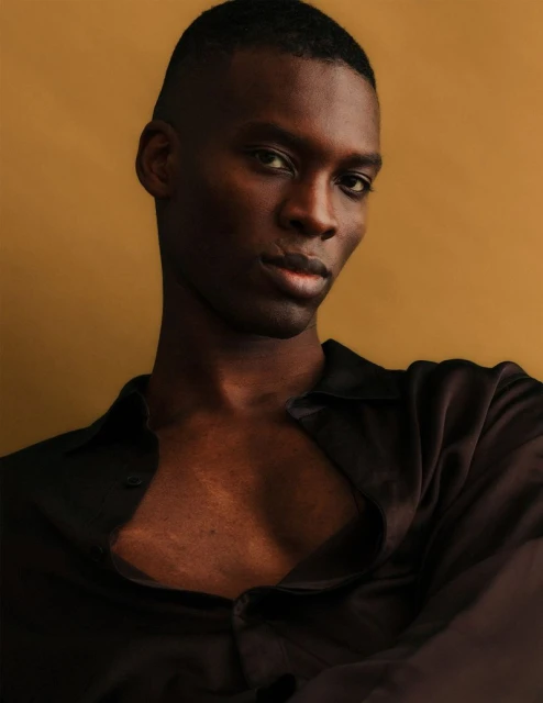 Portrait of an African American male model wearing a black satin button-down shirt against a mustard-yellow background in soft lighting.