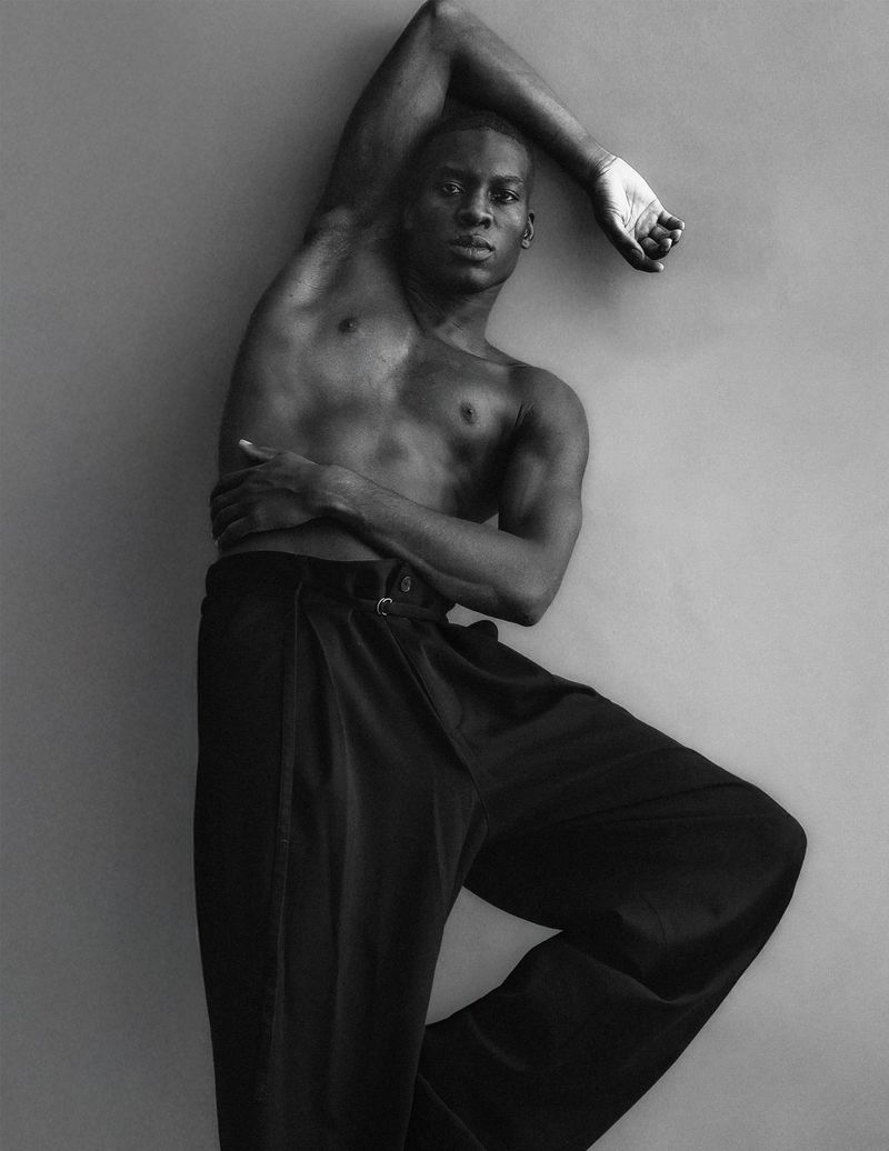 Portrait of an African American man with bare chest wearing black pants in a fashion pose against a grey background in black and white.
