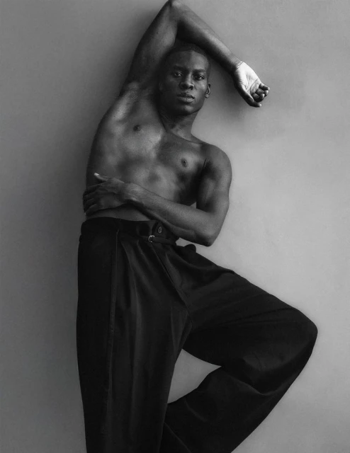 Portrait of an African American man with bare chest wearing black pants in a fashion pose against a grey background in black and white.