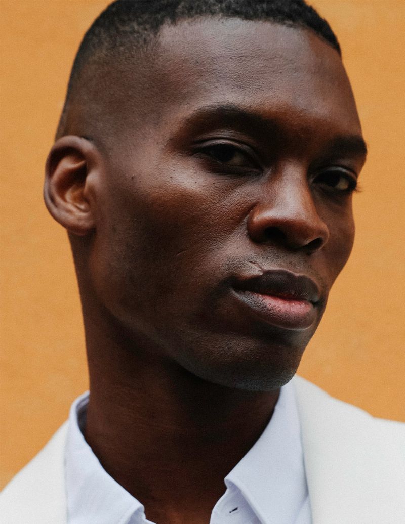 Close-up portrait of a Black man wearing a white collared shirt against a warm ochre background.