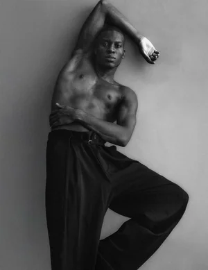 Shirtless Black man posing with raised arm in wide-leg black pleated trousers against a grey backdrop.