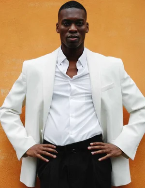 Black man wearing a tailored white blazer and open-collar shirt posing with hands on hips against an orange backdrop.