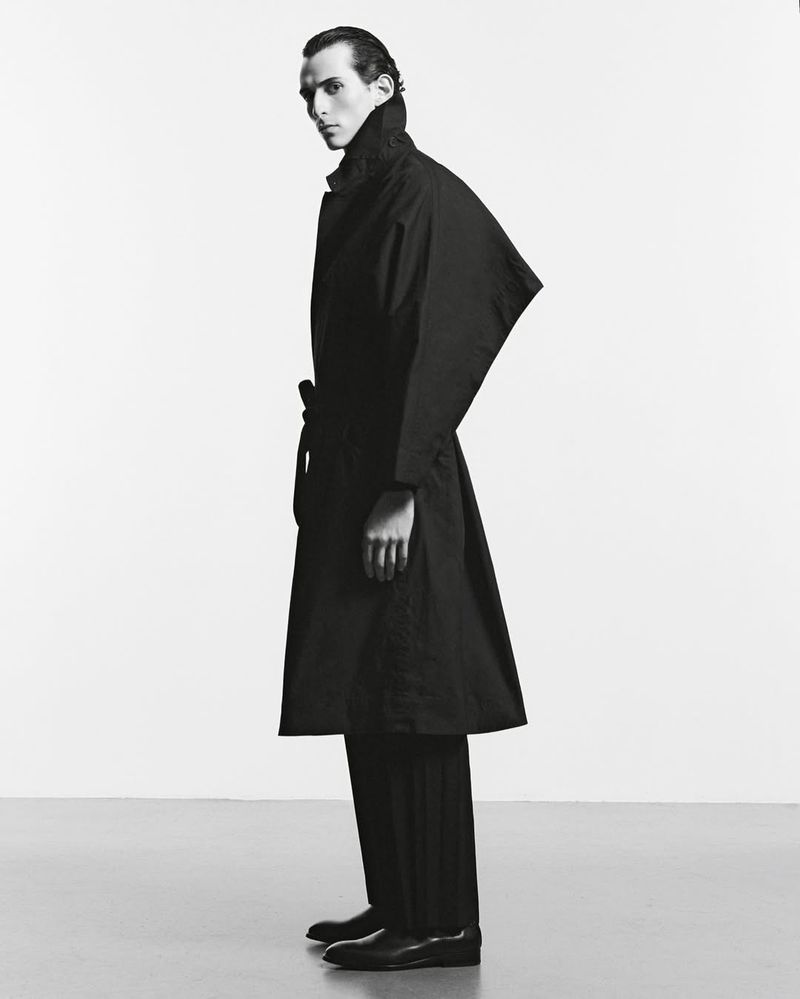 Model Fares Ben M'barka posing in profile wearing a structured black belted coat and dark trousers against a pale studio background.