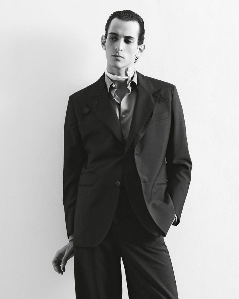 Monochrome portrait of a slender adult male in a dark suit with a scarf, posing indoors against a light wall, exuding a sophisticated, fashion-forward style.