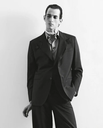 Monochrome portrait of a slender adult male in a dark suit with a scarf, posing indoors against a light wall, exuding a sophisticated, fashion-forward style.