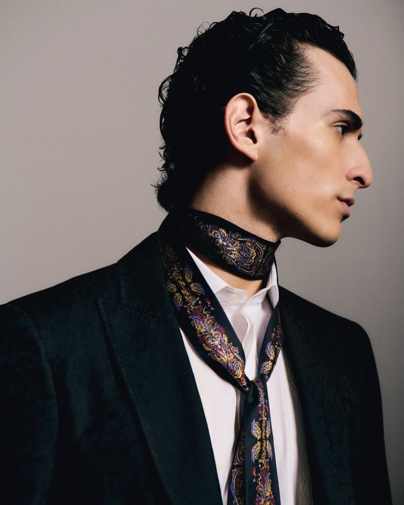 Profile view of a fair-skinned adult male in a black blazer and white shirt, wearing a patterned ascot, against a grey backdrop.