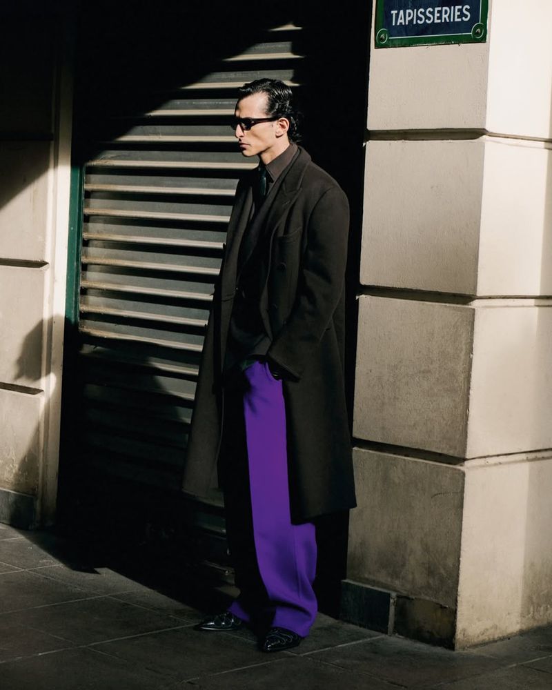 A stylish adult man stands beside a building with purple pants, a dark coat, and sunglasses in an outdoor urban environment.