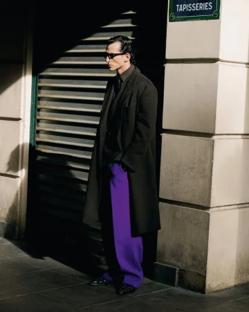 A stylish adult man stands beside a building with purple pants, a dark coat, and sunglasses in an outdoor urban environment.