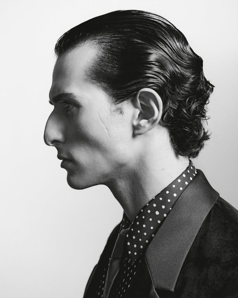 Monochrome portrait of adult male in profile with slicked-back hair, wearing a polka dot shirt, dark suit, and tie, against a plain background.