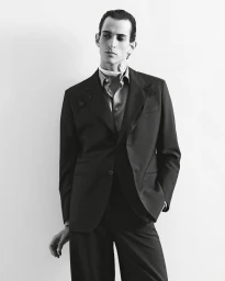 Model Fares Ben M'barka wearing a dark tailored suit and neck scarf posing against a white studio background in black and white.