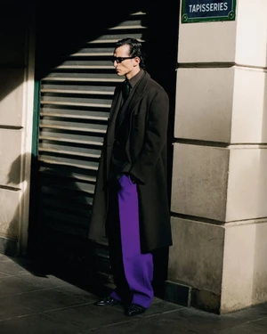 Model wearing a dark tailored overcoat and bright purple trousers standing in high-contrast sunlight on a city street.