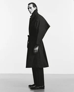 Model Fares Ben M'barka posing in profile wearing a structured black belted coat and dark trousers against a pale studio background.