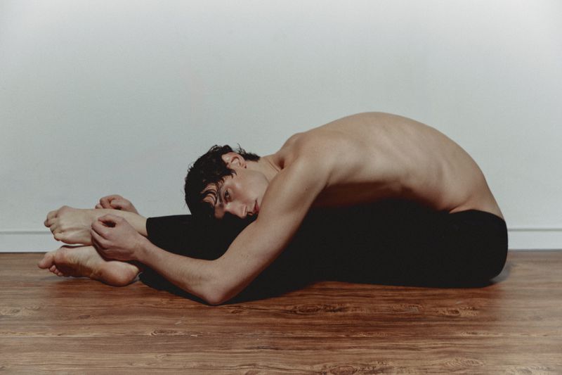 Young adult man lies on wooden floor in full-body stretch against white wall in black athletic pants for fitness and lifestyle imagery.