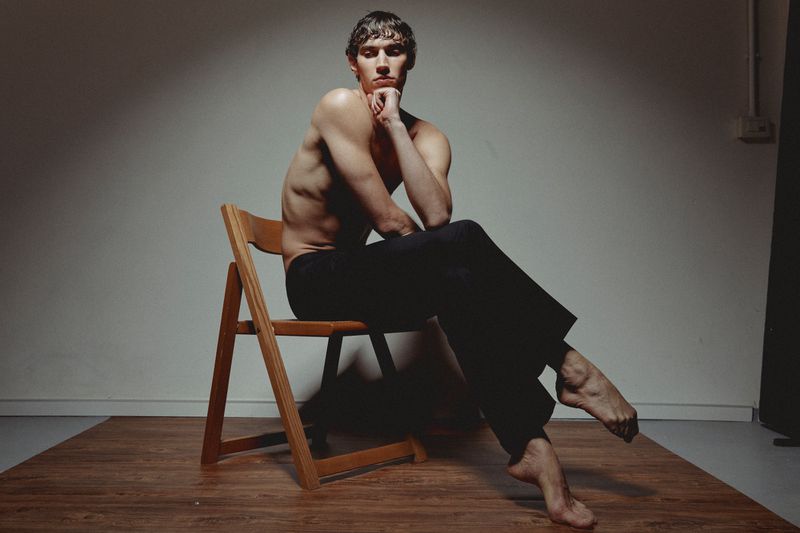 Shirtless man with black pants sits barefoot on wooden chair in photo studio, hand on chin, pensive expression, against grey backdrop.
