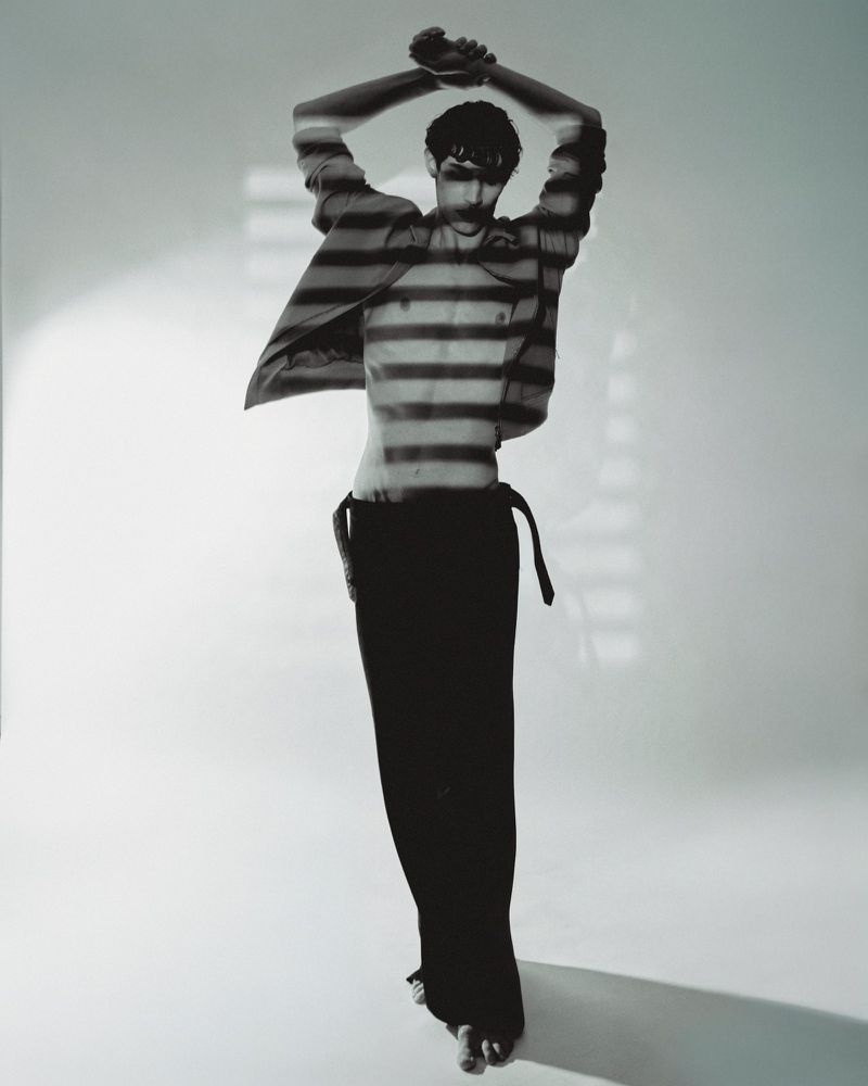Full-length studio shot of an adult male with arms raised, shadowed by horizontal lines against a plain backdrop, wearing an open shirt.