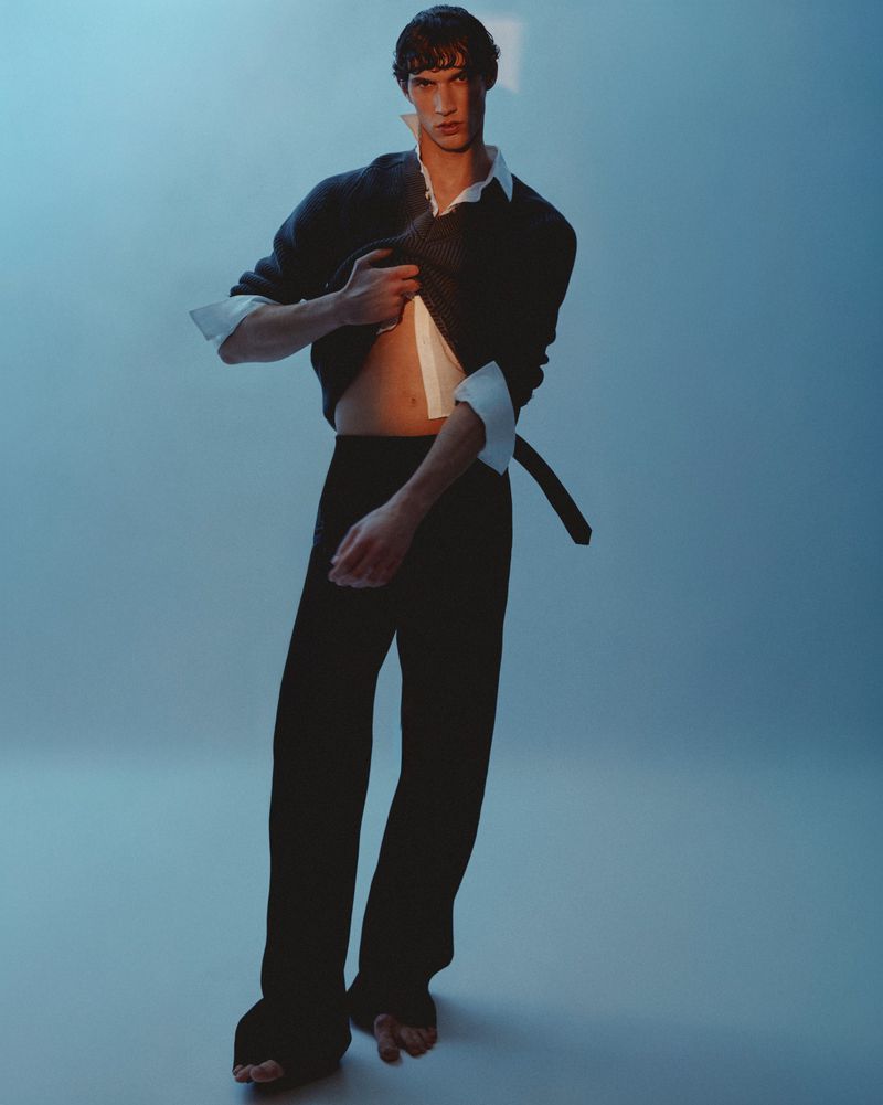 Marco Bozzato posing barefoot in dark trousers and an unbuttoned shirt under a sweater against a cool blue studio backdrop.