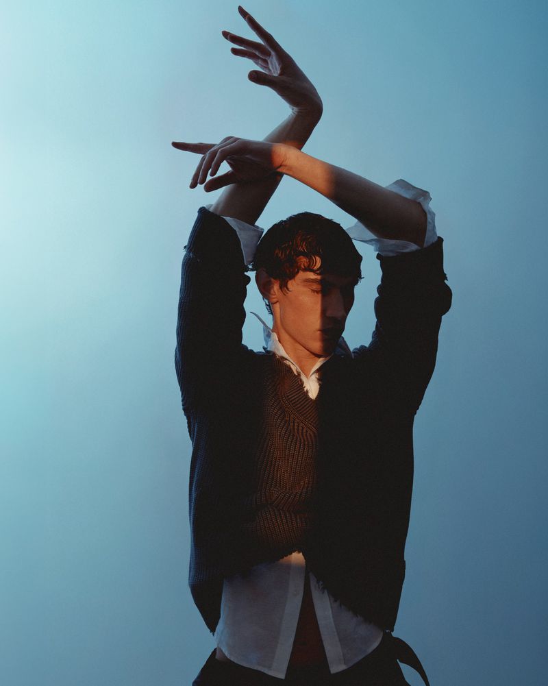 Artistic studio shot of a young adult man posing with arms raised against a gradient blue background, with split lighting and shadow play.