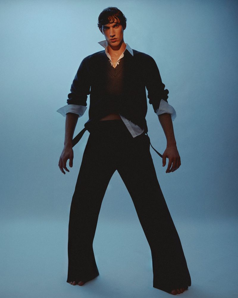 Stylish young adult man in an unbuttoned white shirt, knit vest, and flared black trousers stands barefoot in a light blue studio.