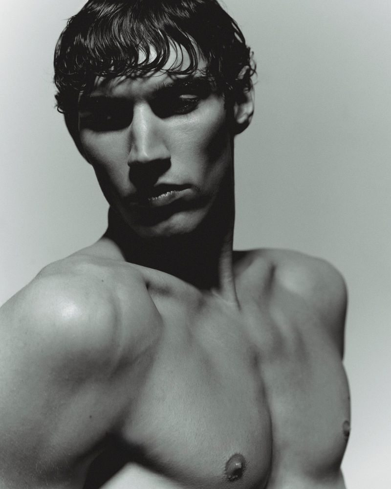 Monochrome portrait of adult male model with dark, wet, wavy hair, smooth skin, and shirtless physique in a studio setting.