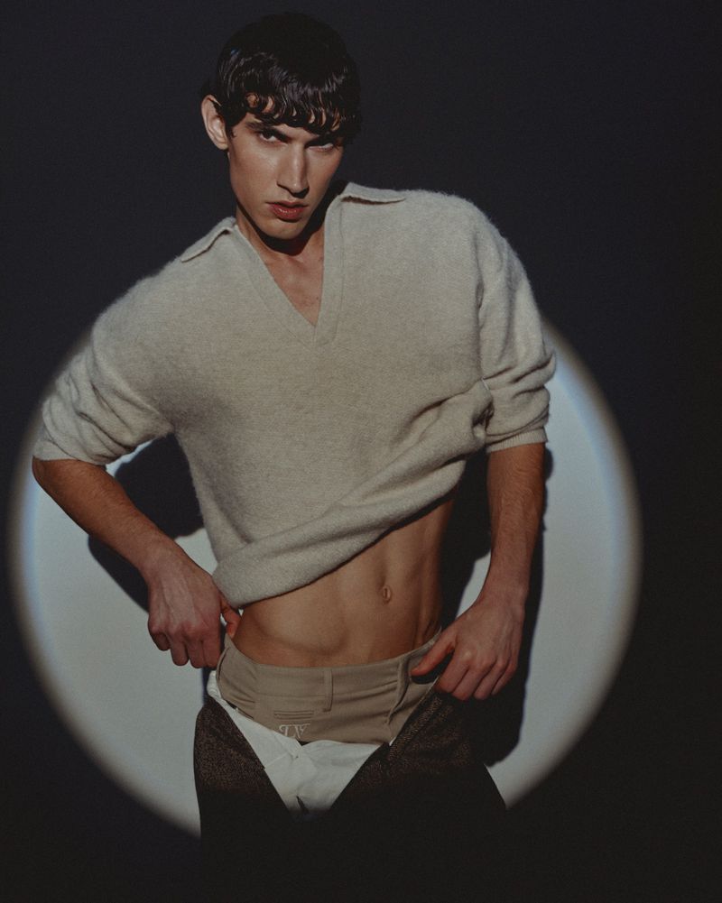 Marco Bozzato posing in a cream knit sweater and layered trousers illuminated by a circular spotlight against a dark background.