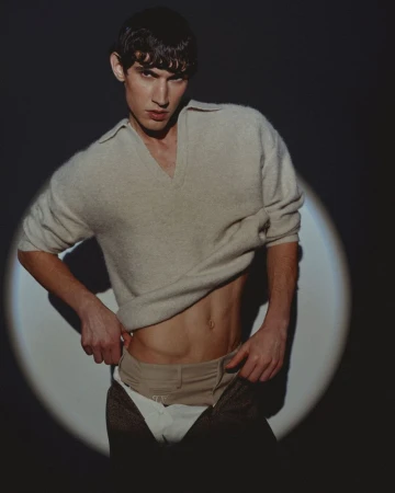 Marco Bozzato posing in a cream knit sweater and layered trousers illuminated by a circular spotlight against a dark background.