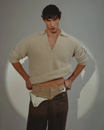 Marco Bozzato posing in a cream knit polo sweater and lowered trousers in a spotlight against a studio backdrop.