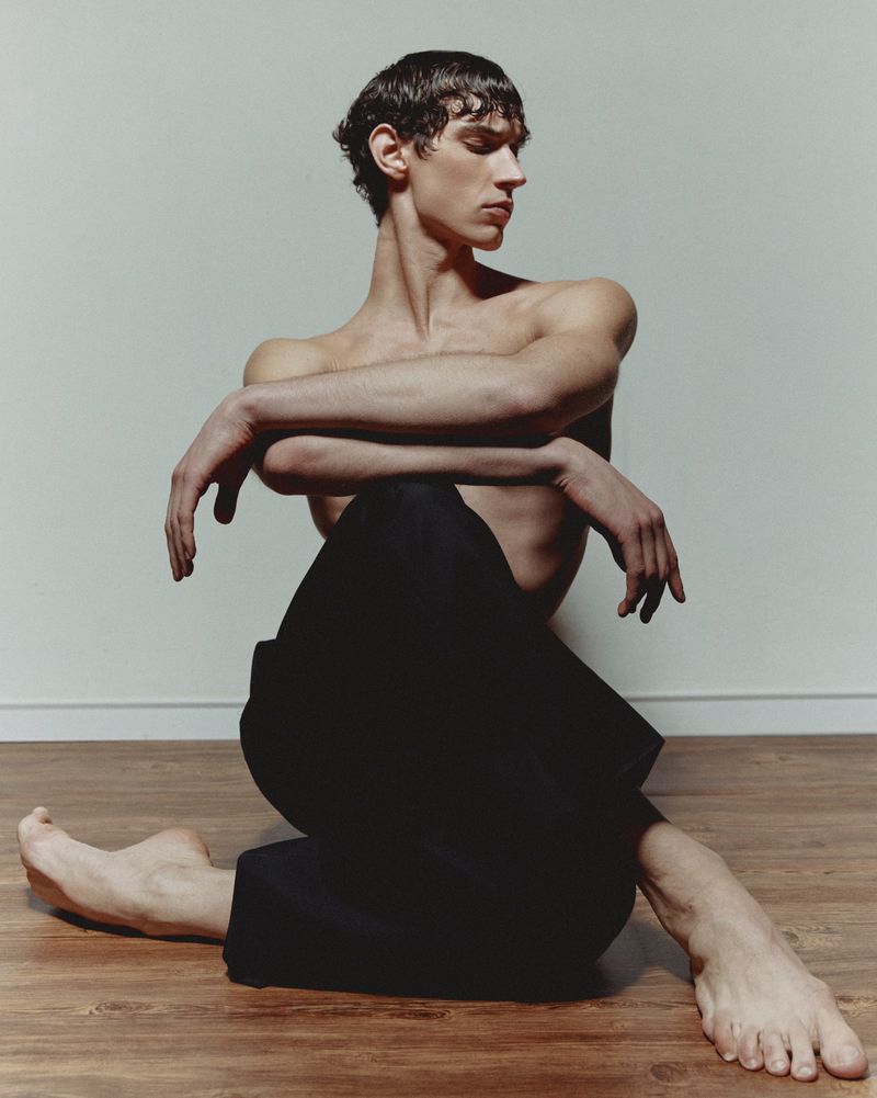 Adult man in a seated pose with bare torso and crossed arms, wearing black pants, against a white wall in an indoor setting.