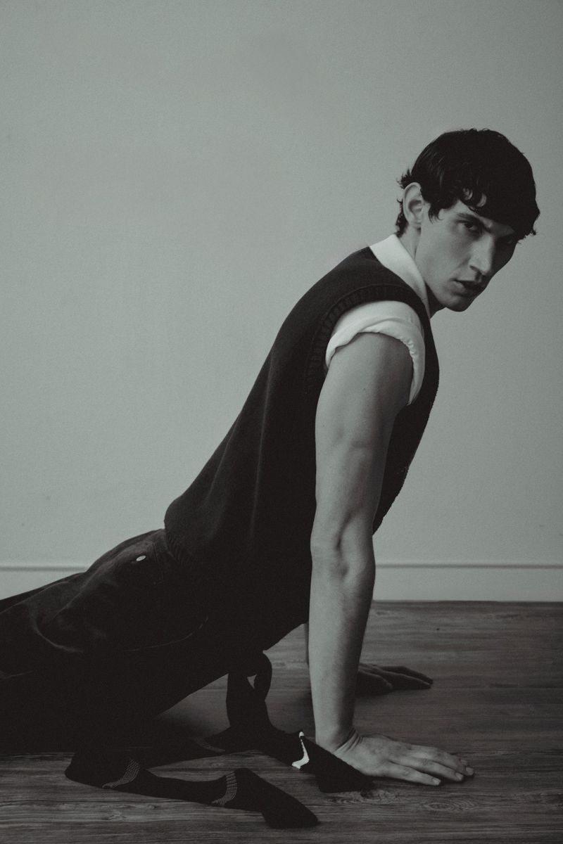 Portrait of a young adult male in a black vest and white shirt, lying on a wooden floor in a minimalist, monochromatic indoor setting.