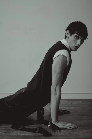 Portrait of a young adult male in a black vest and white shirt, lying on a wooden floor in a minimalist, monochromatic indoor setting.