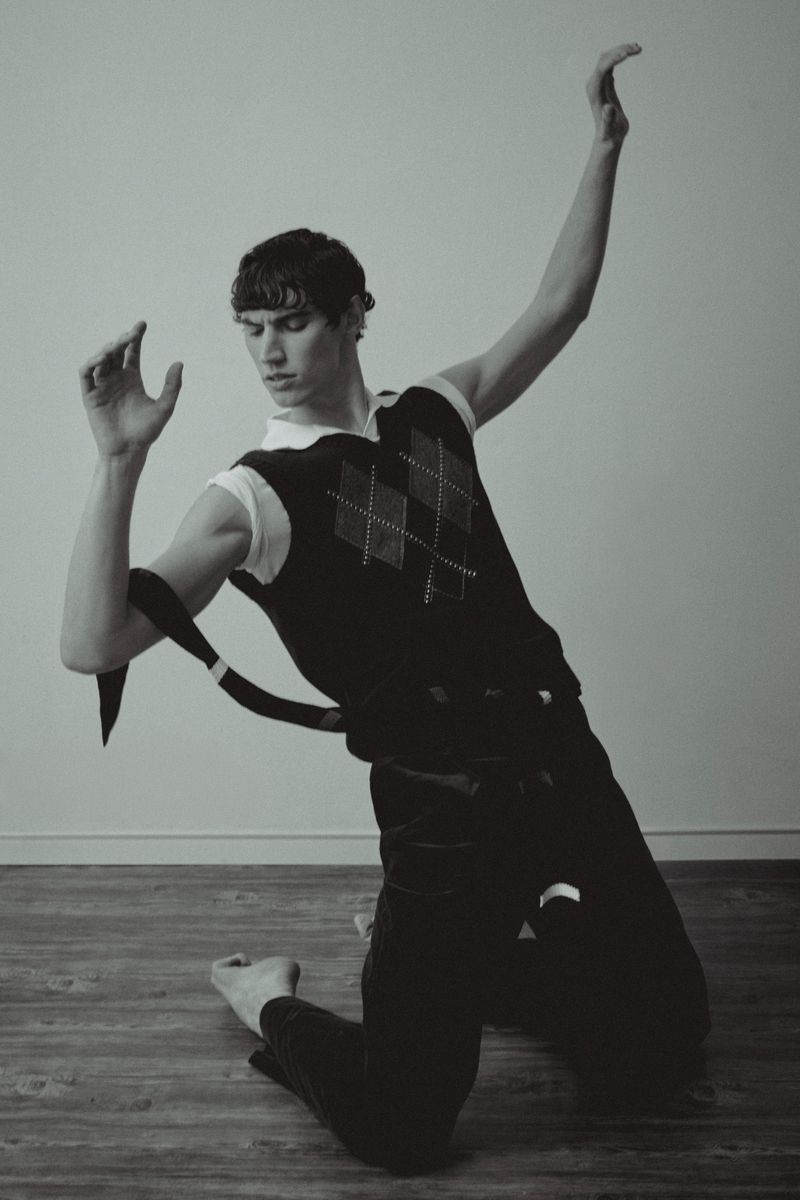 Monochromatic portrait of a young adult male in an argyle sweater vest, kneeling in a dance pose with arms raised in front of a neutral background.