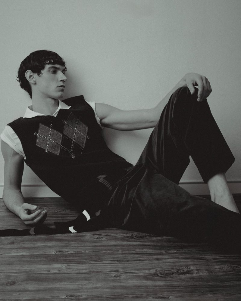 Young man lounges on a wooden floor in a black and white studio shot. He's wearing an argyle sweater vest and dark pants.