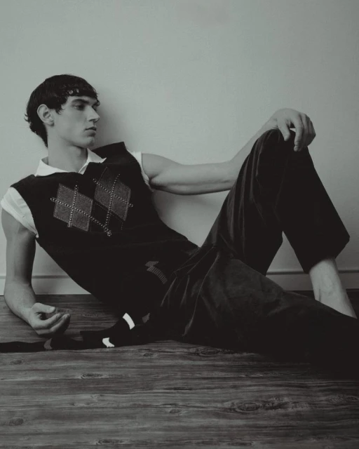 Young man lounges on a wooden floor in a black and white studio shot. He's wearing an argyle sweater vest and dark pants.