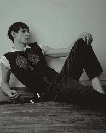Young man lounges on a wooden floor in a black and white studio shot. He's wearing an argyle sweater vest and dark pants.