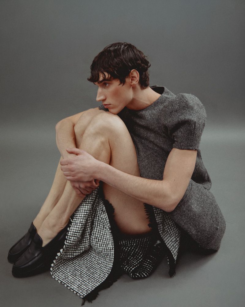 Person sitting in a photo studio with a gray backdrop, wearing a gray top, checkered skirt, and black shoes, facing away from the camera.