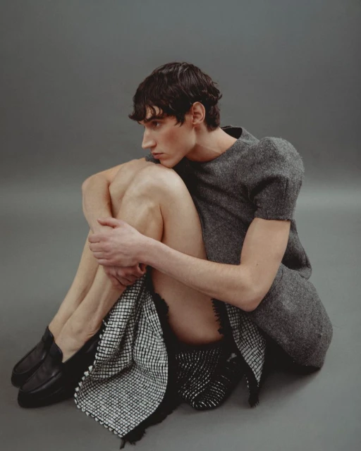 Person sitting in a photo studio with a gray backdrop, wearing a gray top, checkered skirt, and black shoes, facing away from the camera.