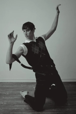 Marco Bozzato kneeling on a wooden floor in an expressive dance pose wearing an argyle sweater vest and dark trousers.