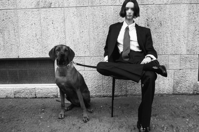 Model in a black tailored suit and tie seated on a chair next to a large dog on a leash against a textured exterior stone wall.