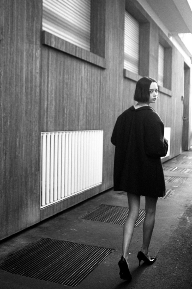 Monochrome portrait of a woman in a dark coat walking away, glancing back over her shoulder in an indoor corridor with window details.