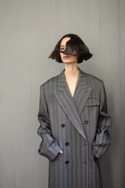 Adult female in a grey striped oversized coat poses in front of a grey background, with short dark hair partially covering her face.