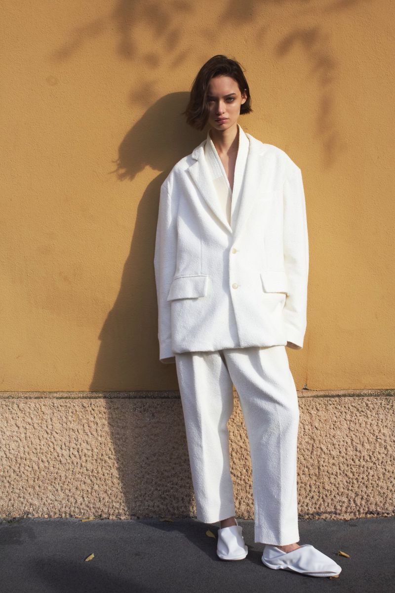 A woman poses against a golden wall, wearing a white blazer, pants, and shoes, with tree shadows adding depth to the fashion portrait.