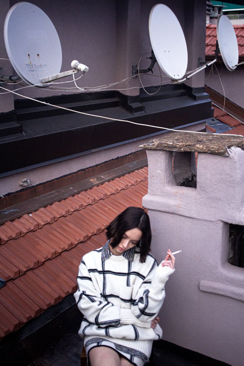 Young woman in a patterned sweater sits on a rooftop with satellite dishes, smoking a cigarette with a city view.