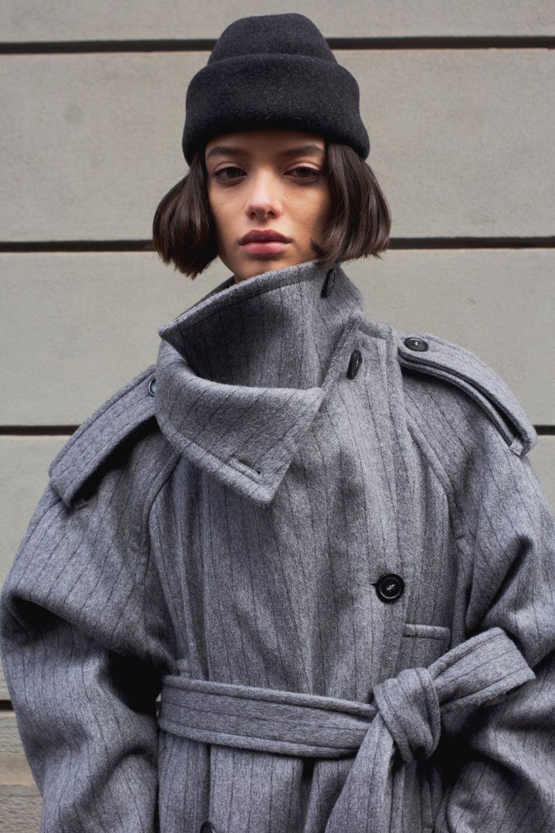 Fashion portrait of a young woman in a grey trench coat and black beanie, standing against a concrete wall in natural light.