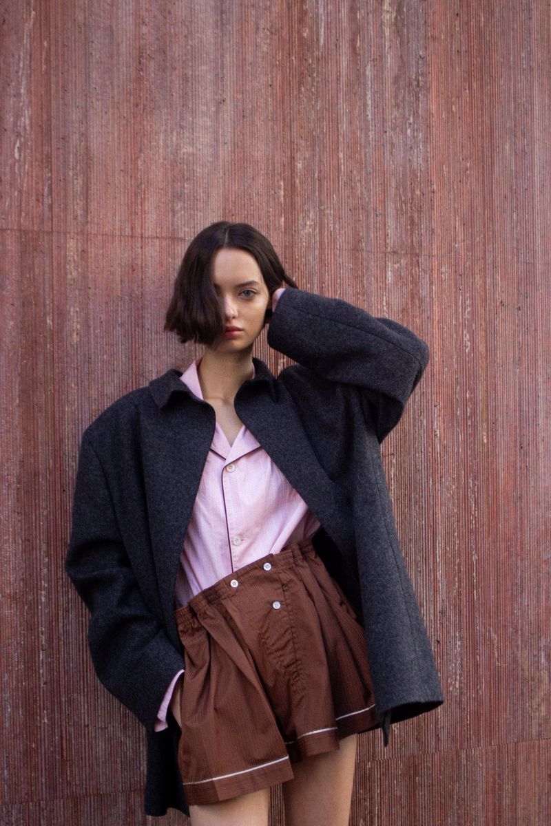 Model Anick Chan wearing an oversized charcoal coat, pink buttoned shirt, and brown pleated shorts against a textured red wall.