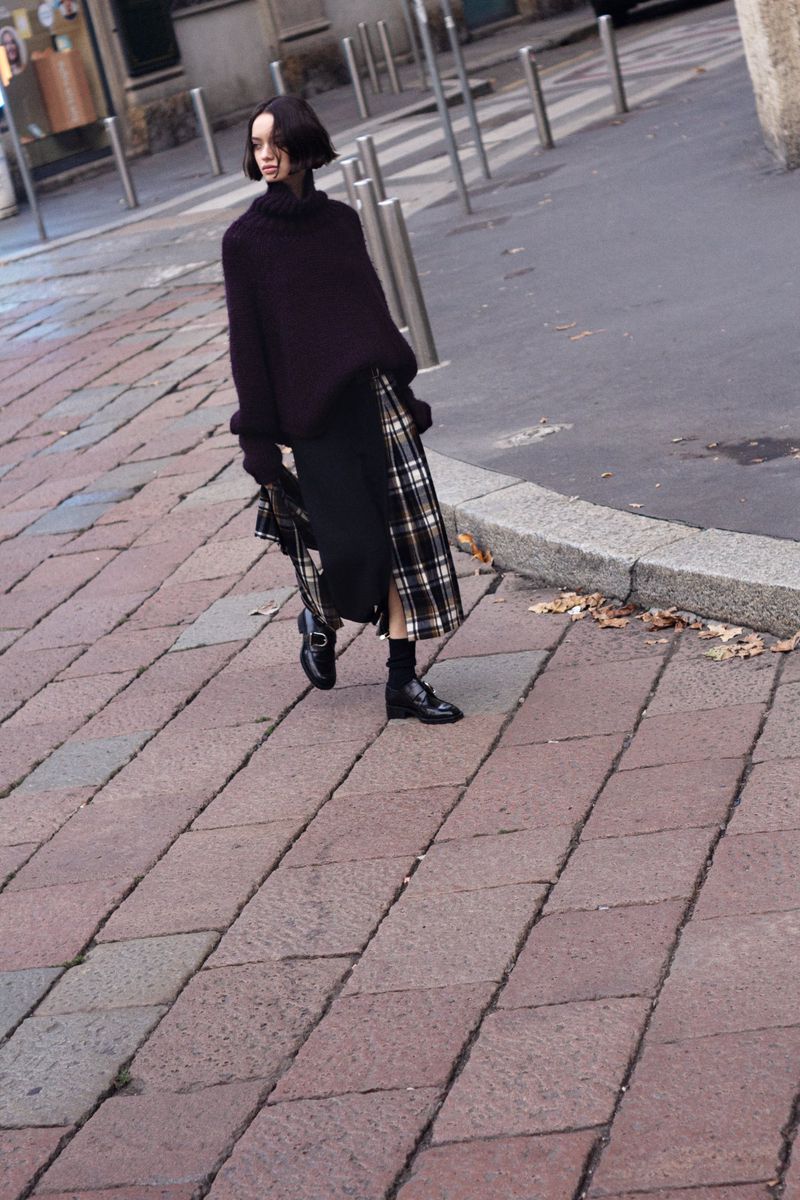 Woman in a turtleneck sweater and plaid skirt walks on a brick sidewalk outdoors with parked cars and buildings in the background.