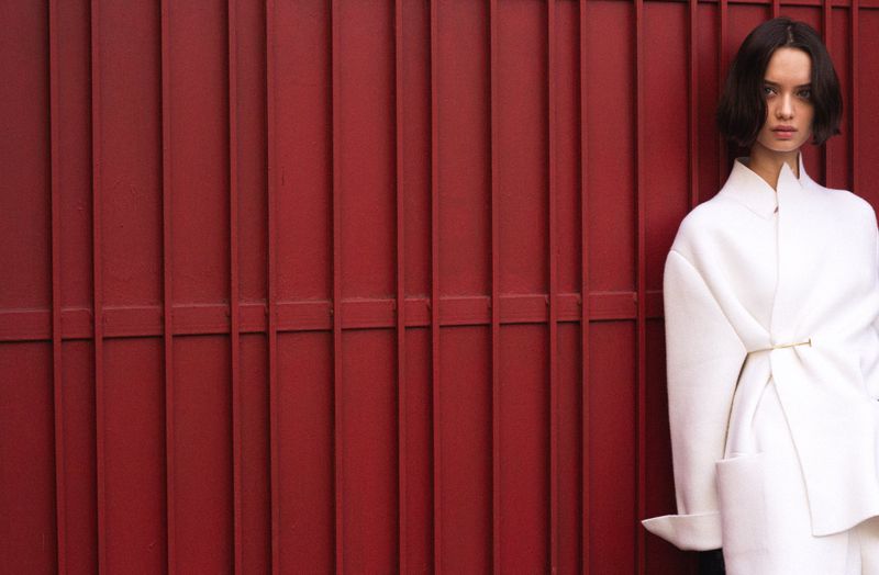 Woman in white coat stands against a red metal fence. Portrait shot showcases fashion against a textured background with natural lighting.