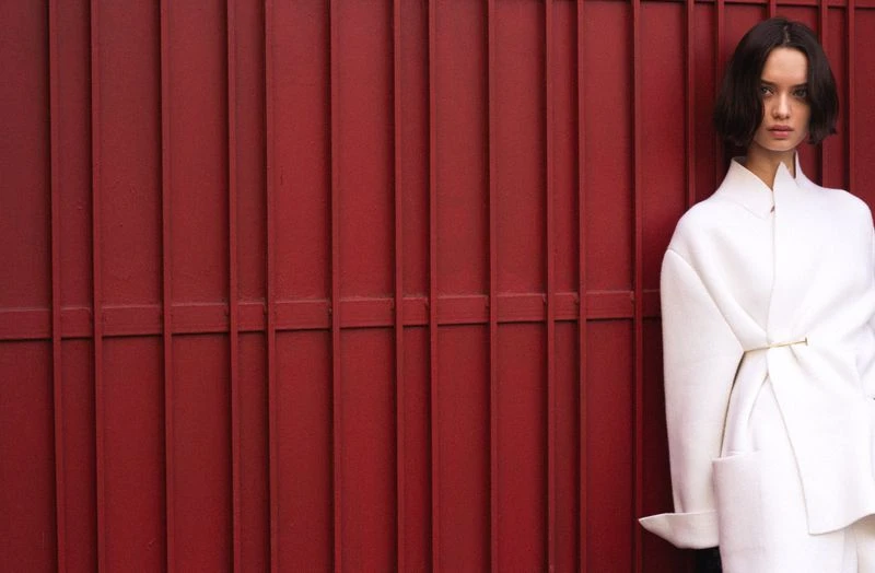 Woman in white coat stands against a red metal fence. Portrait shot showcases fashion against a textured background with natural lighting.