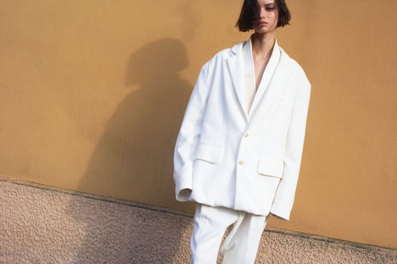 Fashion shot of a young woman in an oversized white suit posing in front of a textured beige wall on an overcast day.