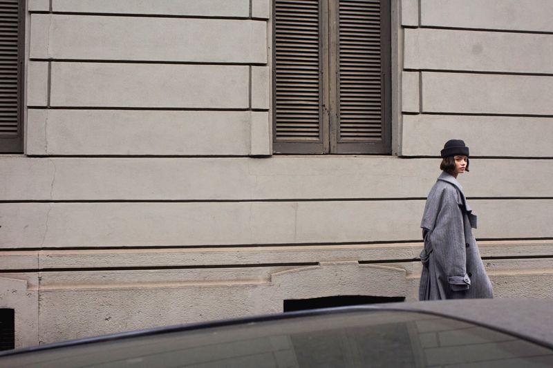 An adult female walks along a city sidewalk in a gray coat and beanie hat. The background features a light gray building with dark gray windows.