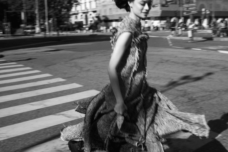 A woman in a textured dress walks across a zebra crossing in a city street, captured in black and white with motion blur.